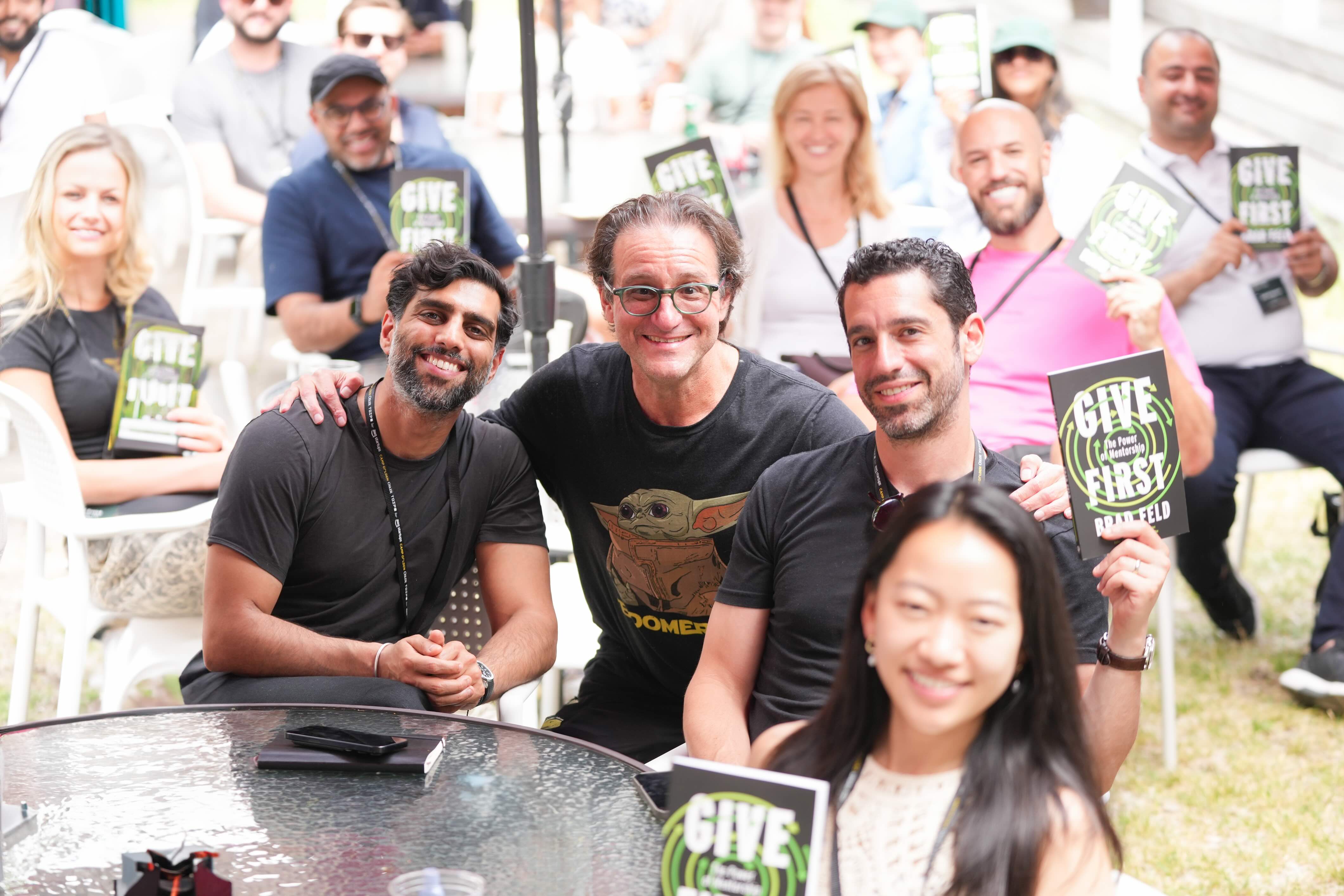 A group of people sitting outdoors, smiling and holding books titled &lsquo;Give First&rsquo; by Brad Feld. In the foreground, two men are posing together with warm expressions, while a woman in front smiles at the camera.