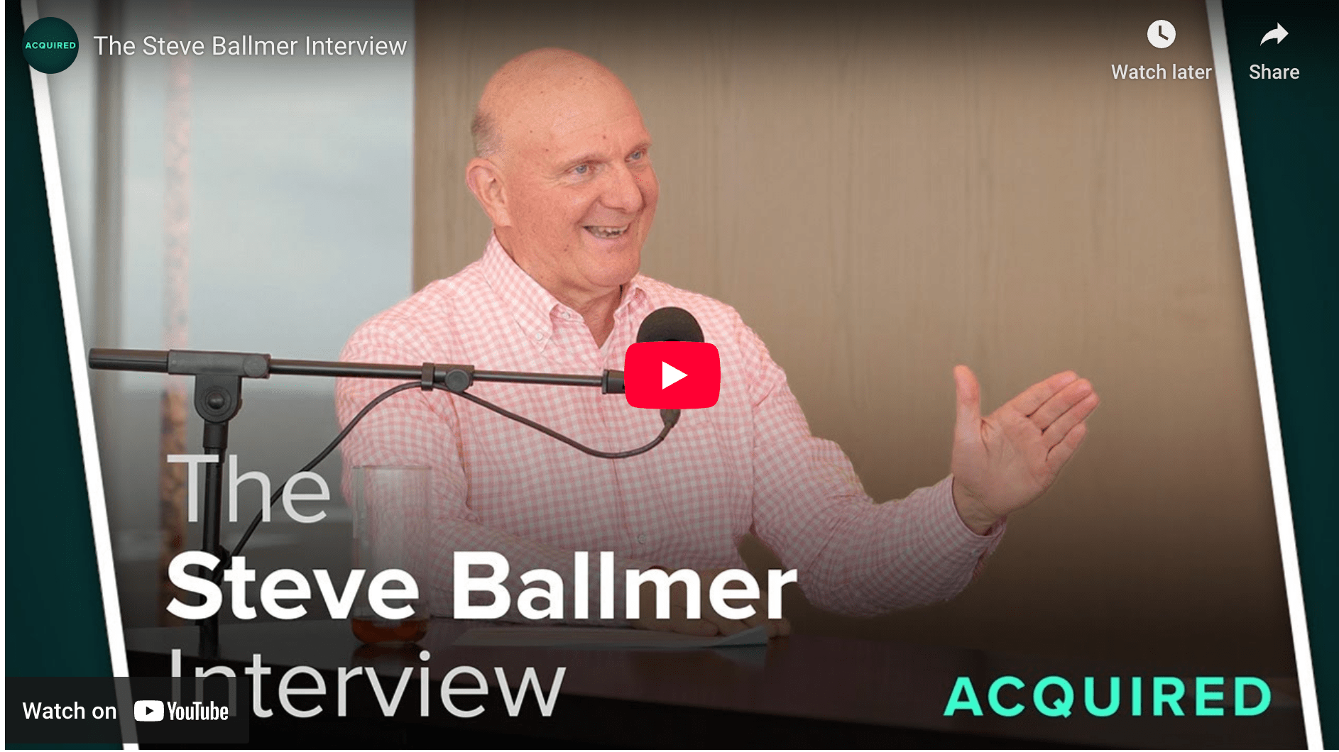 A man with a bald head and a pink checkered shirt gestures while speaking at a microphone during &lsquo;The Steve Ballmer Interview&rsquo; podcast. The background features a beige wall, and a play button is prominently displayed.