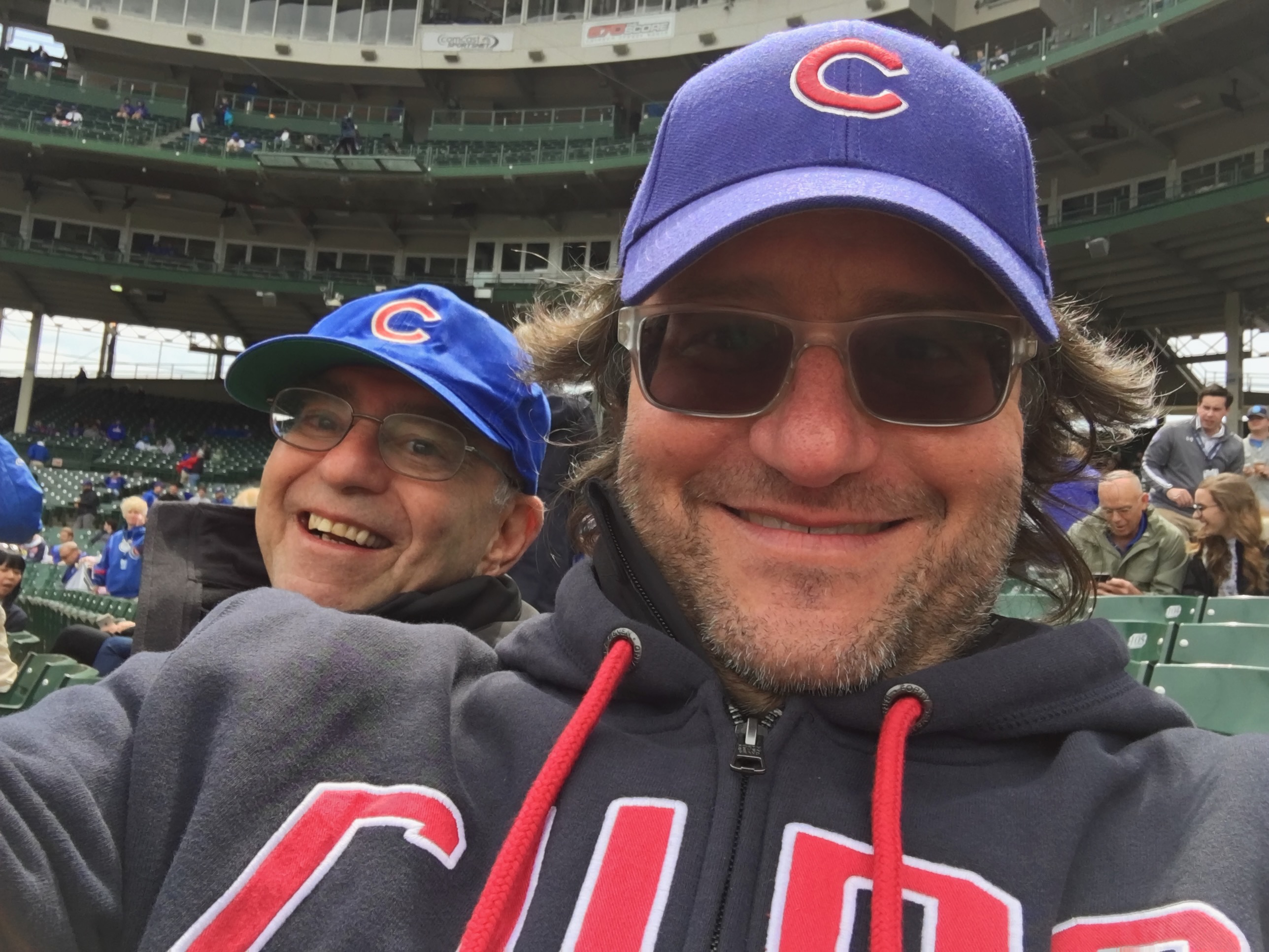 Two men smiling for a selfie while wearing Chicago Cubs hats at a baseball game.