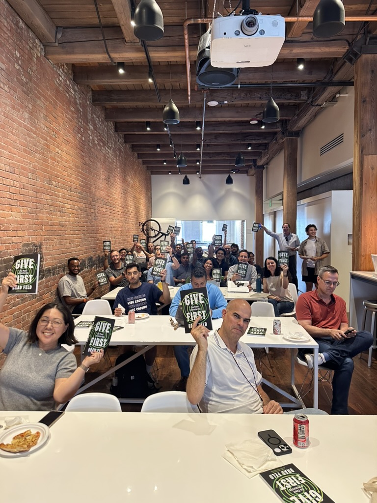 A group of people holding up signs that read &lsquo;Give First&rsquo; in a bright, open room with exposed brick walls and wooden beams.