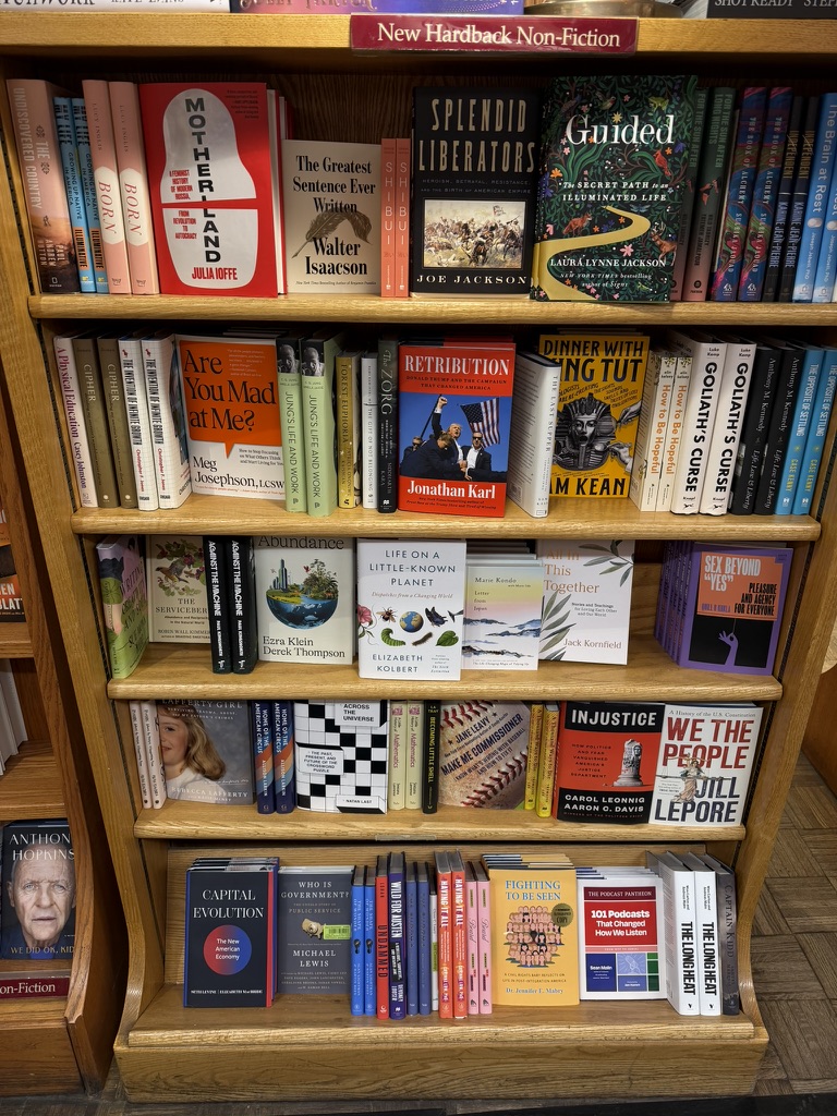 A wooden bookshelf filled with various books, showcasing both fiction and non-fiction titles, including &lsquo;Motherland&rsquo; by Julia Ioffe, &lsquo;Splendid Liberators&rsquo; by Joe Jackson, and &lsquo;Capital Evolution&rsquo; by Michael Lewis.